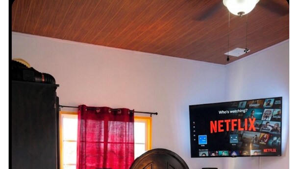 A bedroom with a bed, a TV mounted on the wall displaying Netflix, a window with red curtains, and a bedside table with books. - Welcome to Grace Guest House. A peaceful and comfortable home. (St. Elizabeth Parish)