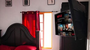 A bedroom with a bed, a window with red curtains, a wall-mounted TV, and a wooden ceiling. - Welcome to Grace Guest House. A peaceful and comfortable home. (St. Elizabeth Parish)
