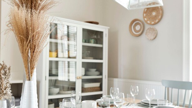 Une table à manger dressée avec des assiettes, des bols et des verres, un vase contenant des plantes séchées et, au fond, un meuble blanc.