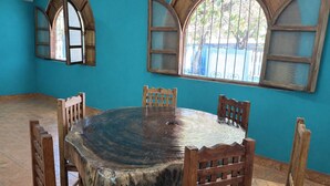A dining area with a round table, four wooden chairs, two arched windows with open shutters, and a tiled floor. - Casa Cruz (Brisas de Zicatela)