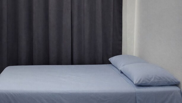 A bed with a grey sheet and a single light blue pillow against a dark grey curtain backdrop. - Casa Cruz (Brisas de Zicatela)