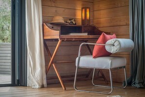 Ein gemütliches Zimmer mit Holzvertäfelung, ein stuhl mit Metallgestell und Sitzkissen, ein kleiner Schreibtisch mit Hut und einem Vintage-Radio sowie ein Fenster mit Vorhängen.