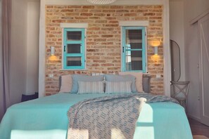 Un dormitorio con pared de ladrillo, dos ventanas con contraventanas azules, una cama con colcha azul y blanca y dos lámparas fijadas a la pared.