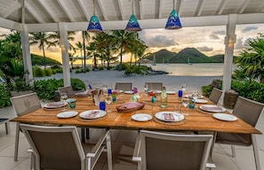 A wooden table set for dining with chairs, a view of the sea, palm trees, and mountains in the background.