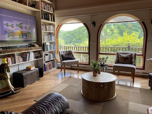 Ein gemütliches Wohnzimmer mit Bücherregal, bequemen Sitzmöglichkeiten, einem runden Couchtisch und großen Fenstern mit Blick auf Grünflächen.
