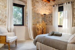 Ein Schlafzimmer mit Steinwänden, einem Bett mit grauer Bettdecke, einem weißen Stuhl und einer Stehlampe.
