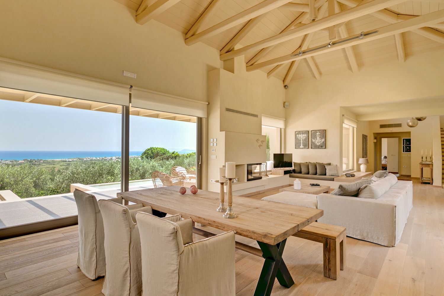 A spacious living area with a wooden table, white sofas, and a large window overlooking a scenic view.