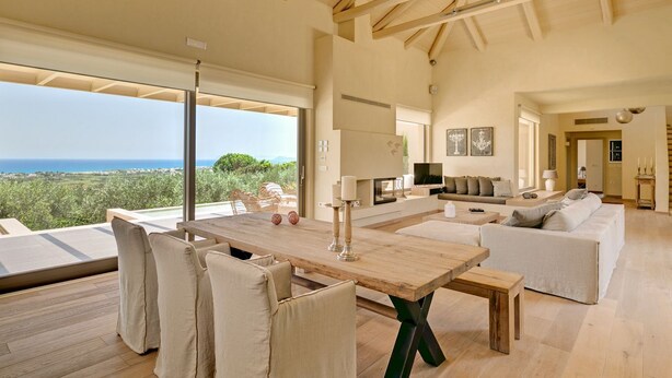 A spacious living area with a wooden table, white sofas, and a large window overlooking a scenic view. - Villa Marat - Four Bedroom Villa, Sleeps 8 (Astratigos)