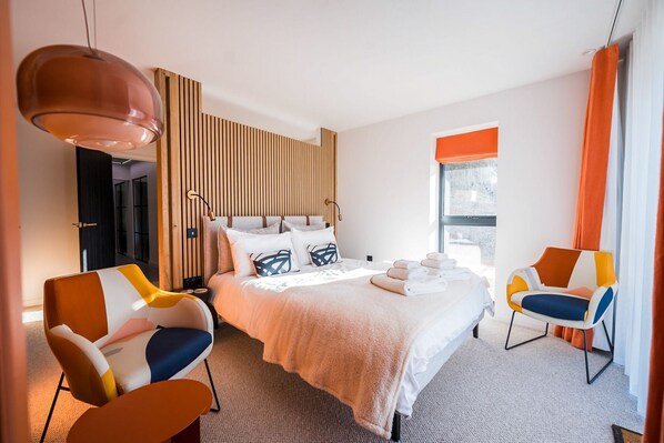 Una habitación de hotel moderna con una cama grande, dos sillas, un panel de pared de madera y una ventana con cortinas.