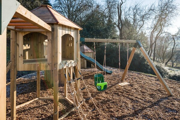 A wooden play structure with a slide and swing set, surrounded by mulch and trees.
