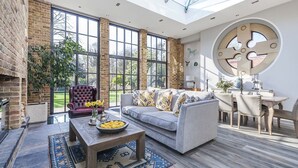 A spacious living area with a large window, a fireplace, a sofa, a dining table, and a circular wall decoration. - Greenwich Hall - Nine Bedroom Villa, Sleeps 22 (London)