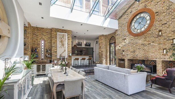 A spacious living area with a high ceiling, a fireplace, and a kitchen in the background. - Greenwich Hall - Nine Bedroom Villa, Sleeps 22 (London)