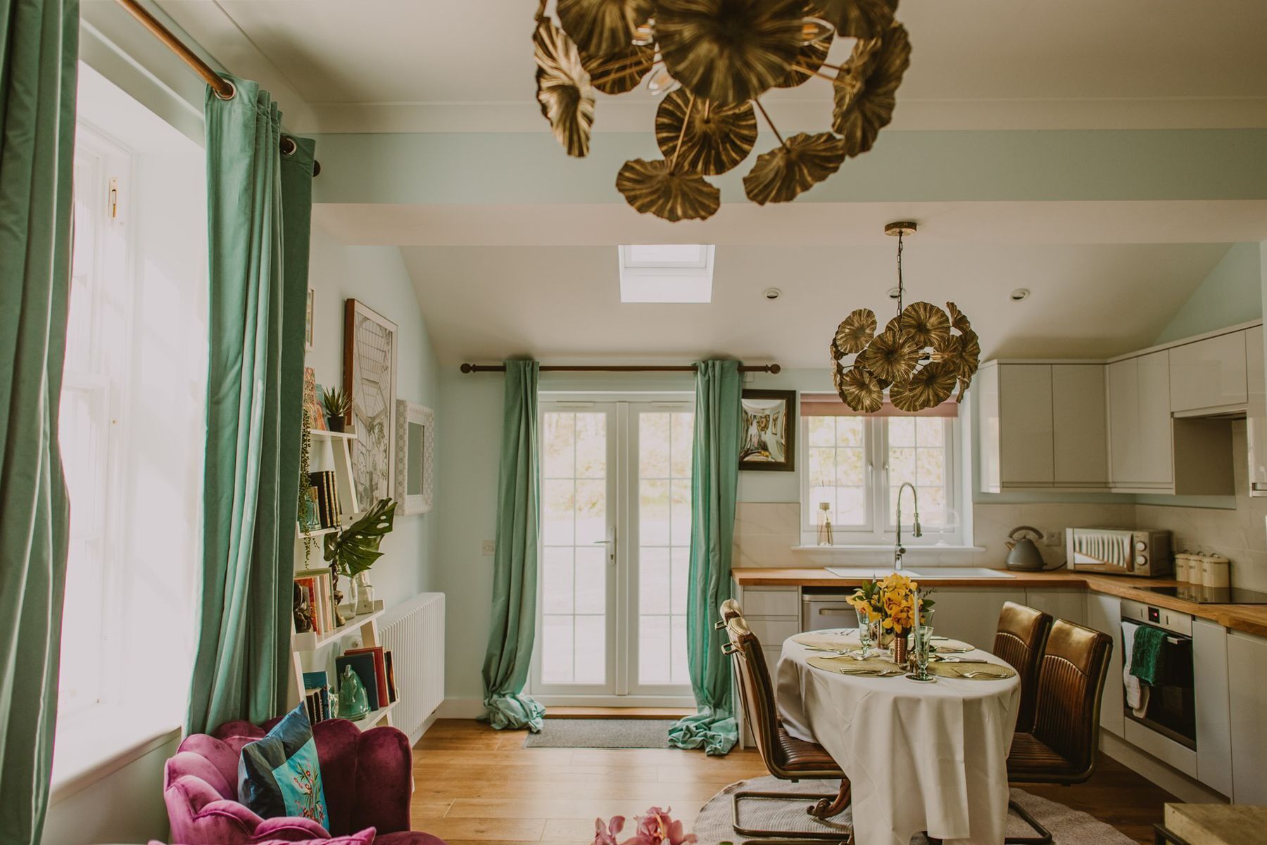 A room with a dining table set for two, a kitchen with white cabinets, and a large window with green curtains.