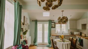 A room with a dining table set for two, a kitchen with white cabinets, and a large window with green curtains.