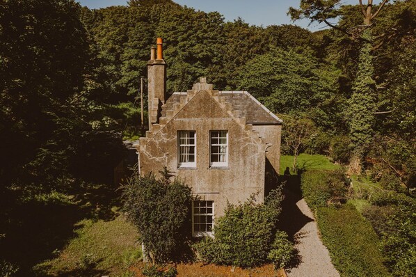 Una casa de piedra con chimenea, rodeada de abundante vegetación y un sendero que lleva a ella.