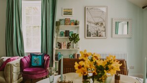 Uma sala de estar aconchegante com um sofá, uma mesa posta para refeições, uma estante com diversos objetos e uma janela com cortinas.