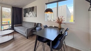 Una sala de estar moderna con sofá, mesa de comedor, sillas, una lámpara colgante y un ventanal con vistas a un edificio.