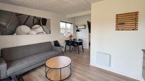 Una sala de estar moderna con un sofá gris, una mesa de centro redonda de madera, un comedor con una mesa y sillas, y un ventanal grande.