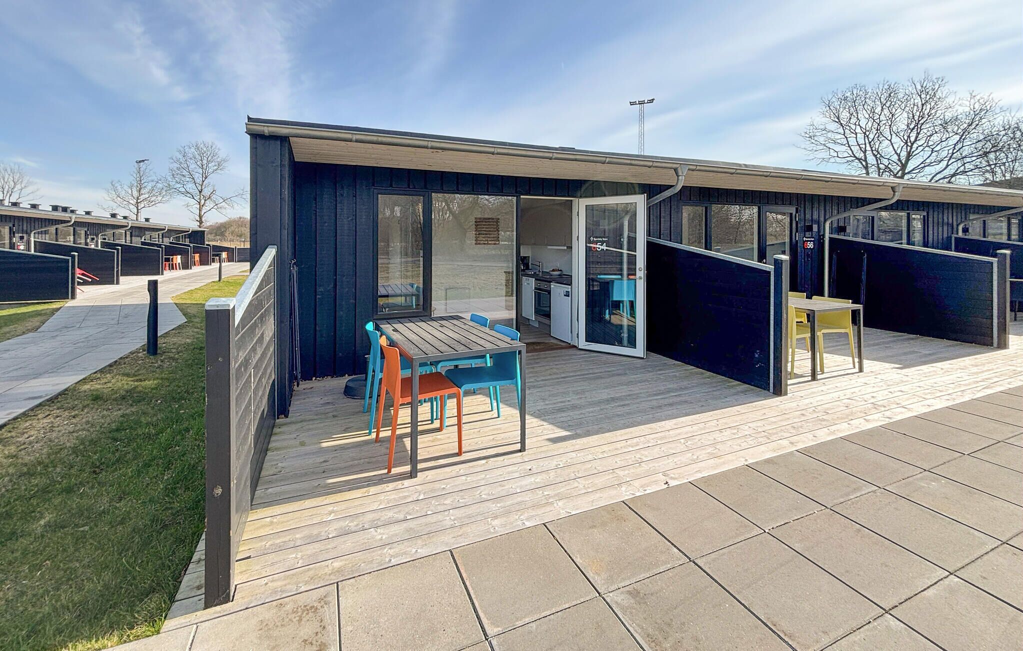 Een modern, compact gebouw met een terras, compleet met houten tafel en stoelen, een glazen deuropening en een kleine keuken die zichtbaar is door het glas.