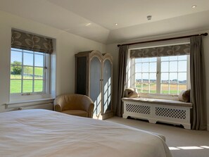 A bedroom with a large bed, a chair, a wardrobe, a window seat, and a view of the countryside.