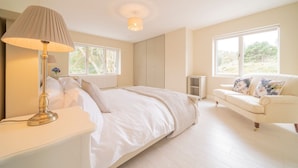 Un dormitorio con cama, lámpara de noche, paredes blancas, ventanas amplias y un sofá blanco.