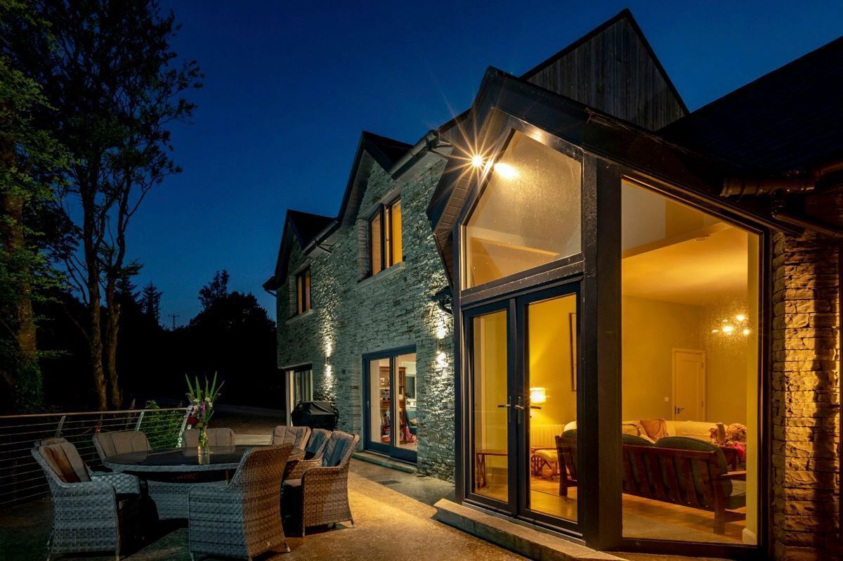 A house with a stone exterior, a large glass door leading to an interior with a bed and seating area, and a patio with outdoor furniture.