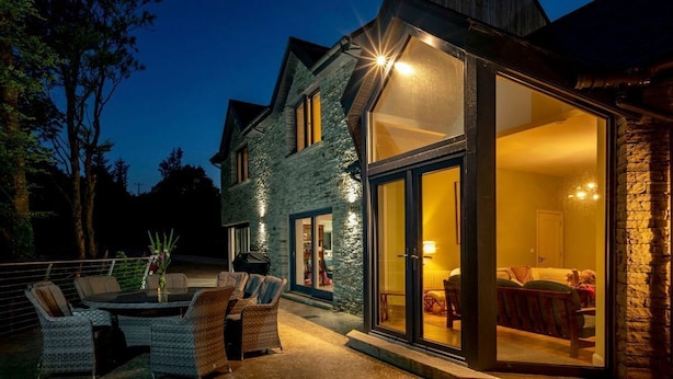 Una casa con fachada de piedra, un gran ventanal que da a un interior con cama y zona de estar, y un patio con mobiliario exterior.