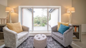 Una sala de estar con dos sillones, un reposapiés redondo y un amplio ventanal corredera que da a un espacio exterior.