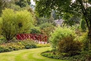 Un puente rojo sobre un sendero curvo en un jardín exuberante con diversos árboles y arbustos.