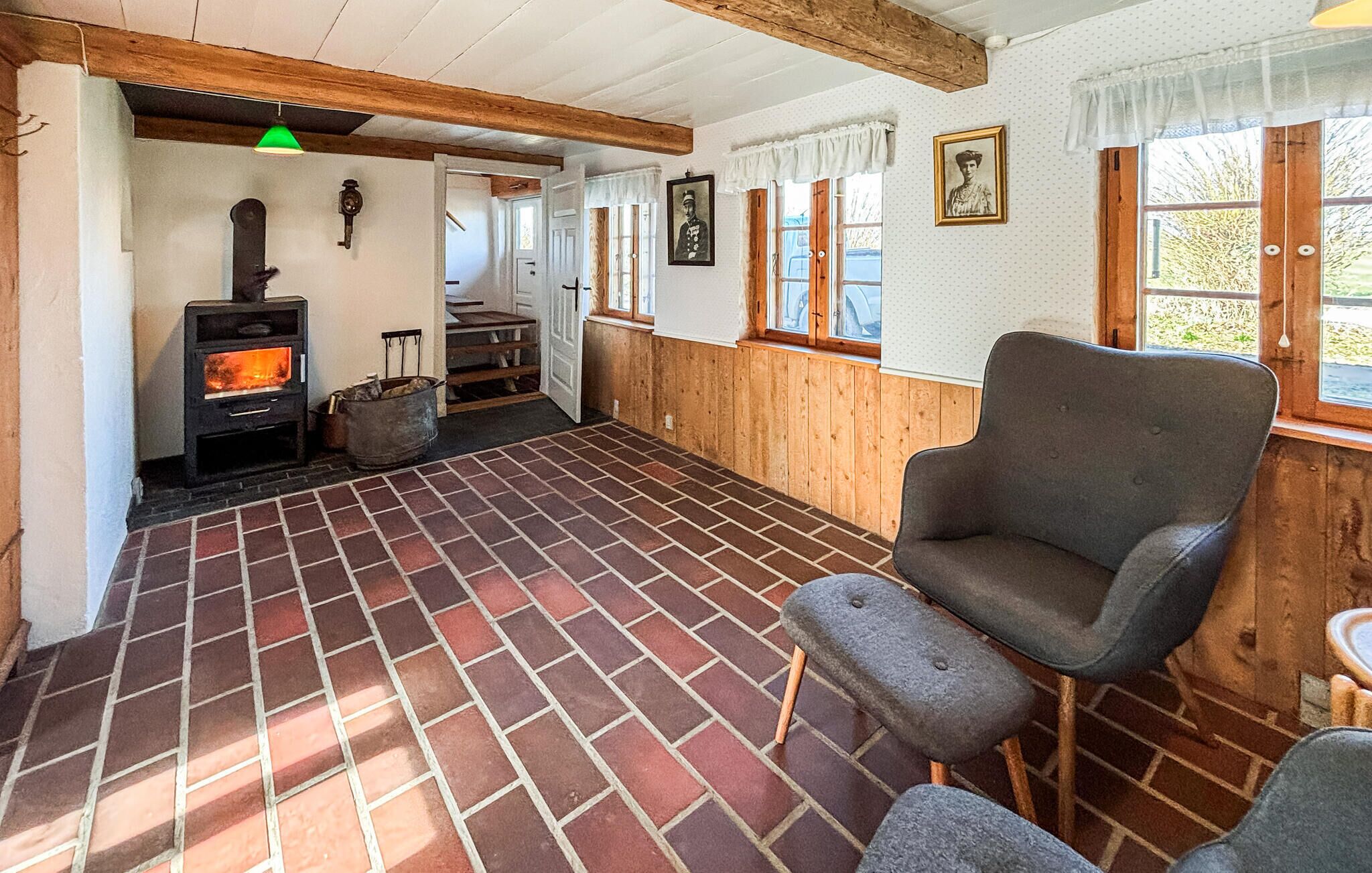 A rustic interior with a brick floor, wooden walls, a stone fireplace, a grey armchair, and a window with a view.