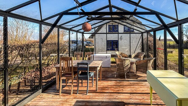 Ein Gewächshaus mit einer Holzterrasse, einem Tisch, Stühlen und einem hängenden Laternen.