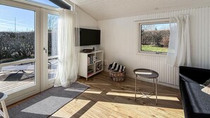 Ein Zimmer mit einer großen Schiebeglasdoor, einem Fernseher auf einem weißen Regal, einem kleinen runden Tisch und Blick ins Freie.