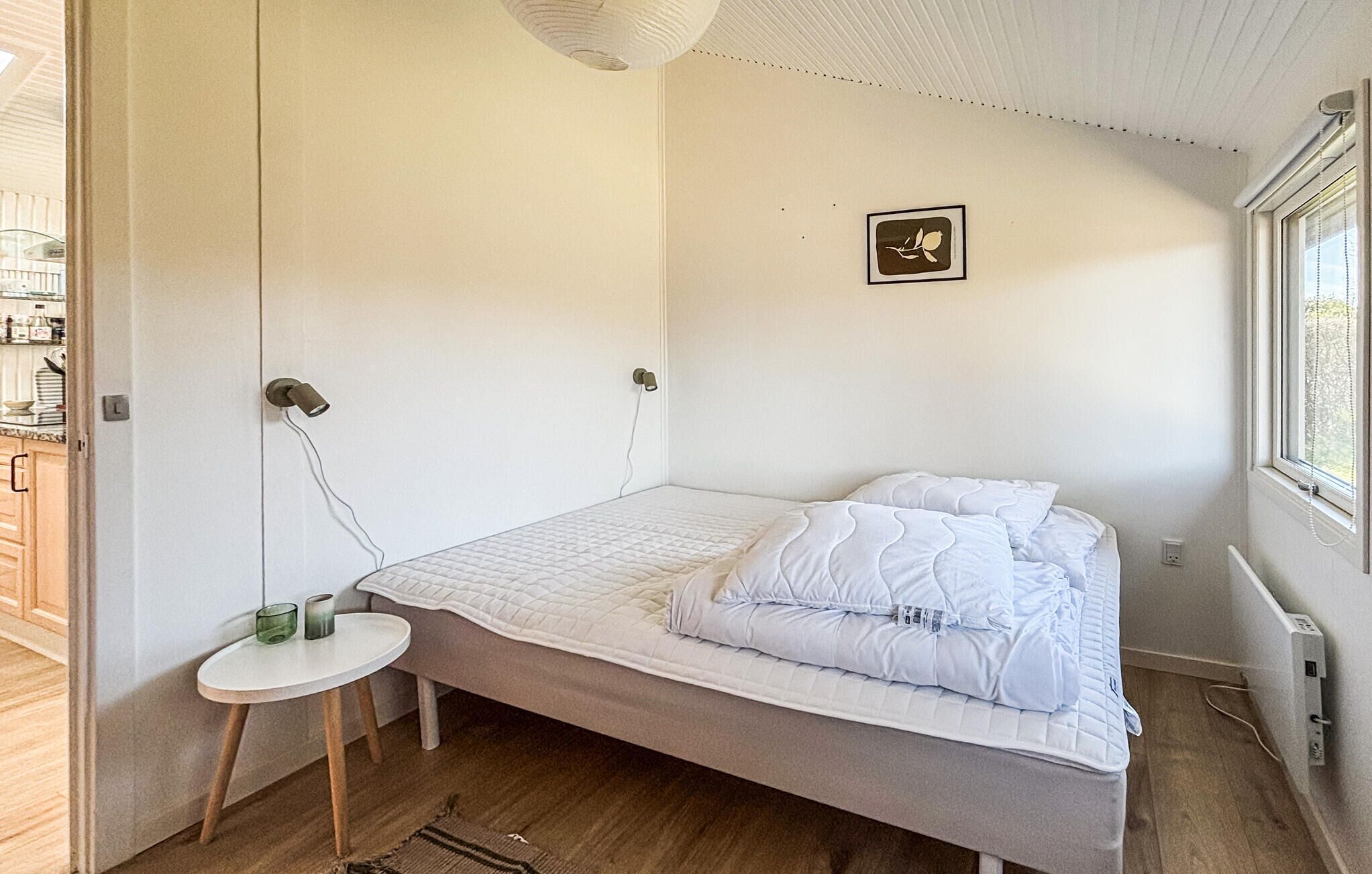 Een enkel bed met witte beddengoed, een klein rond tafeltje met een lamp en een foto aan de muur.