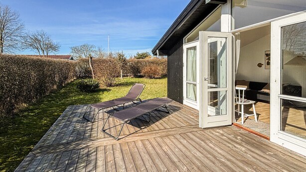 Eine Holzterrasse mit zwei Liegestühlen, eine Glastür zu einem Raum mit einer Couch und einem kleinen Tisch, und ein Hecken im Hintergrund.