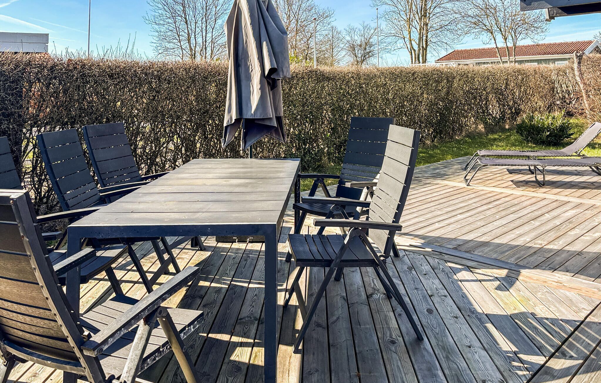 Eine Holzterrasse mit Tisch und Stühlen, einem Liegestuhl und einer Hecke im Hintergrund.