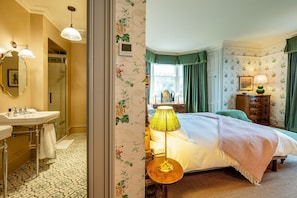 Une chambre d’hôtel avec salle de bain, un lit recouvert d’une courtepointe verte, une table de chevet avec une lampe et une commode.