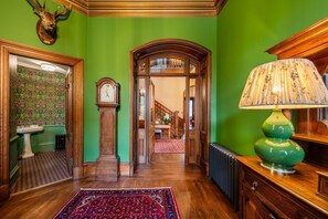Un pasillo con pared verde, suelo de madera, alfombra estampada, un reloj y una lámpara.