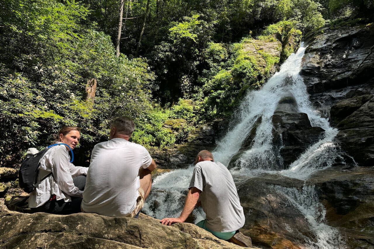 Trois personnes sont assises sur des rochers, près d’une cascade, dans une zone boisée.