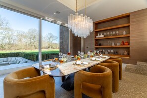Une salle à manger moderne avec une grande table dressée pour six personnes, entourée de fauteuils confortables, d’un lustre et offrant une vue sur l’extérieur.