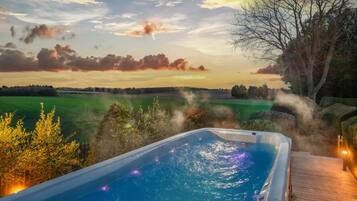 A hot tub with a wooden deck, surrounded by greenery and a view of a field.