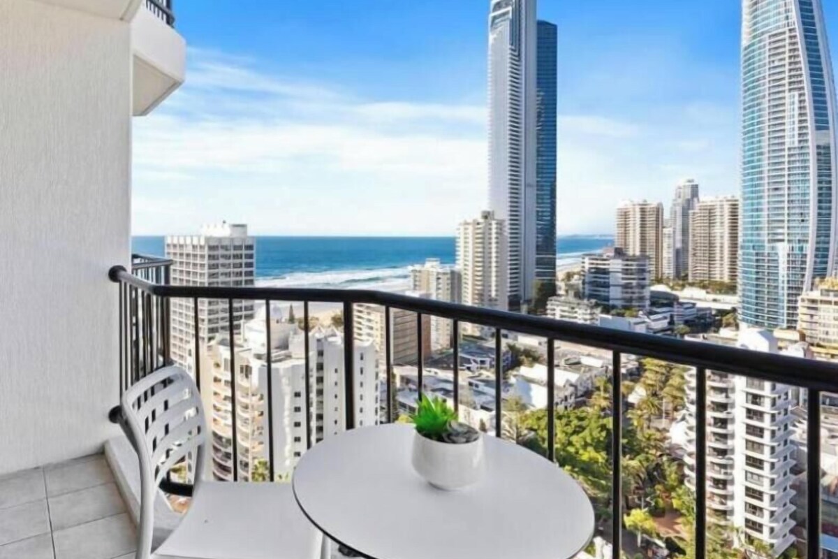 Ein Balkon mit weißem Tisch und Stuhl, einem Blumentopf und Blick auf die Stadt mit hohen Gebäuden und dem Meer.