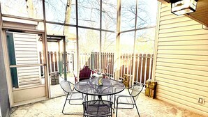 Un patio techado con una mesa redonda de metal y sillas, suelo de piedra, una valla de madera y una casa al fondo.
