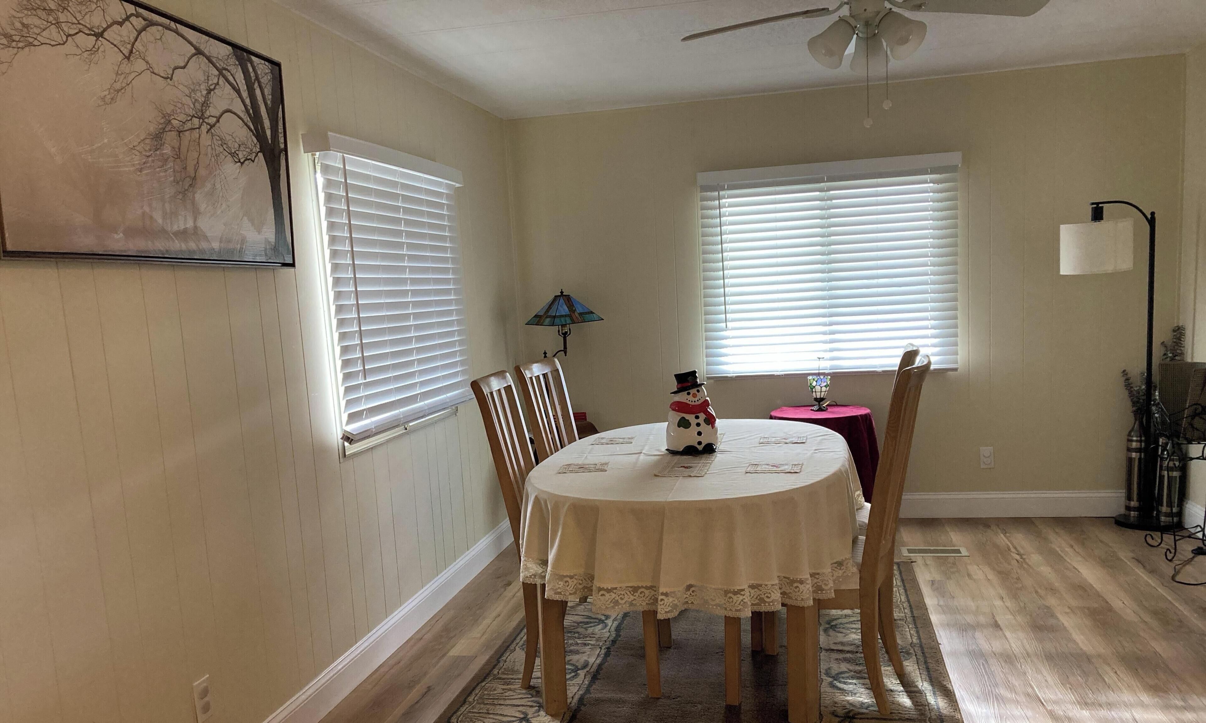 A dining area with a round table, chairs, a ceiling fan, and two windows with blinds.