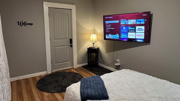 A hotel room with a TV displaying a channel guide, a white door, a black rug, and a white bed with a blue pillow. - Cozy Apartment Little Rock Pets Welcome (Little Rock)