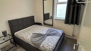 Ein Schlafzimmer mit Bett, Nachttisch, Spiegel, Fenster mit Vorhängen und einer Pendelleuchte.