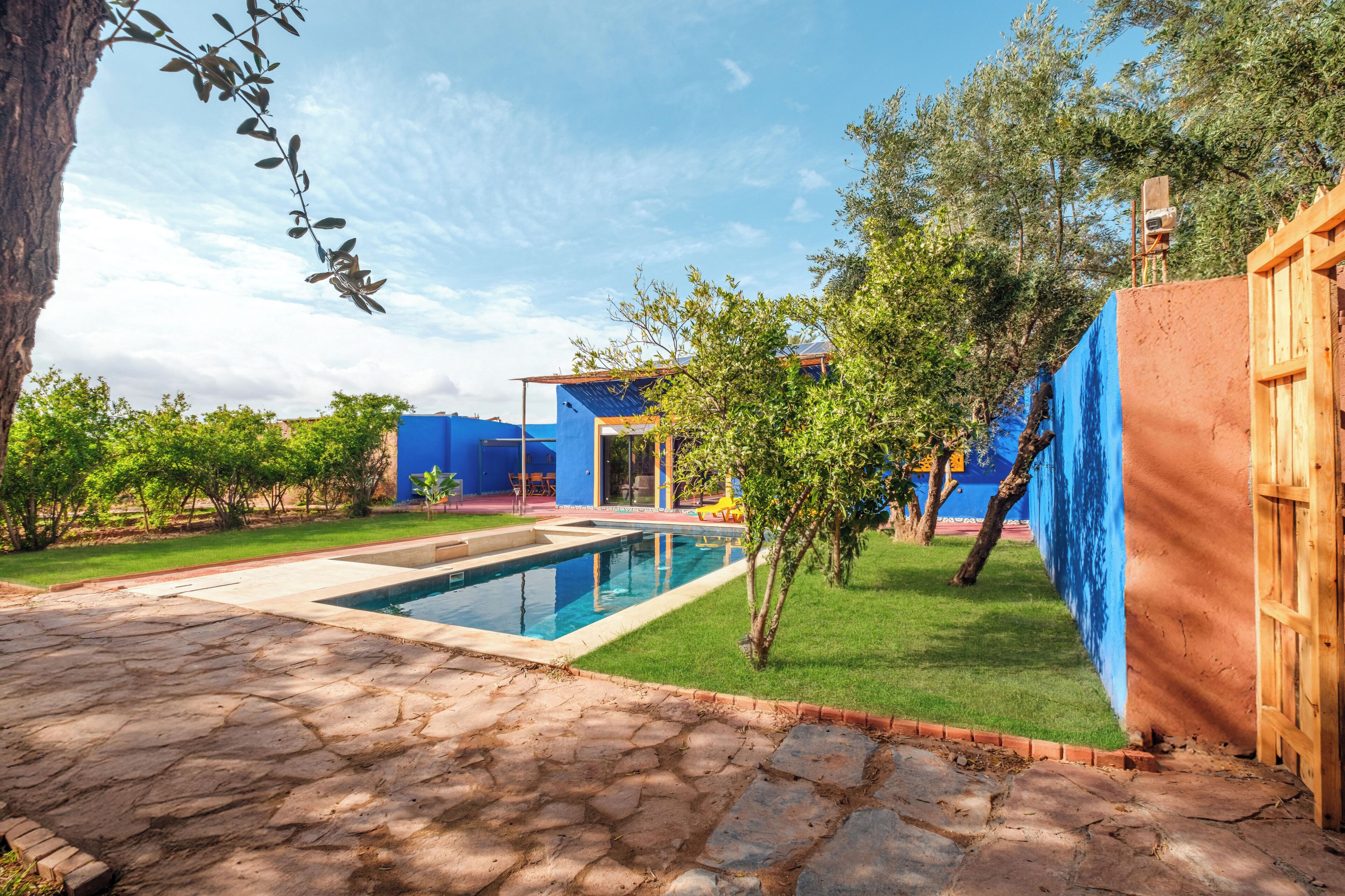 A swimming pool with a stone patio, a blue house, and a wooden fence.