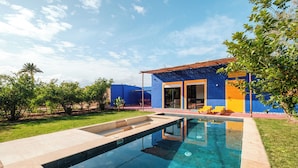 Una piscina con superficie de agua azul cristalina, rodeada por un césped cuidado y una casa con paredes exteriores azules y amarillas.