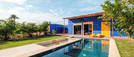A swimming pool with a clear blue water surface, surrounded by a well-maintained lawn and a house with blue and yellow exterior walls.