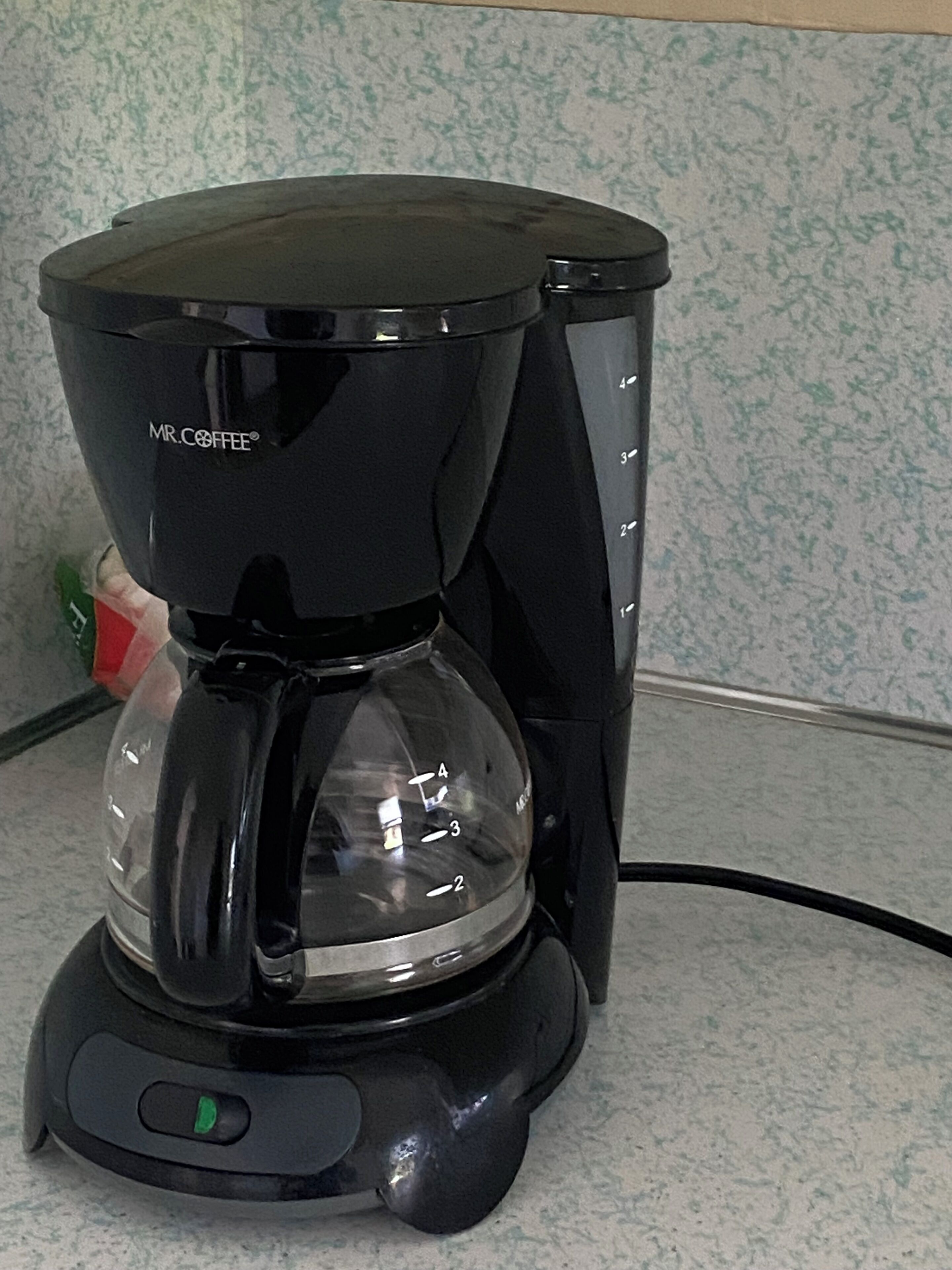 Una cafetera Mr. Coffee negra amb un recipient de vidre transparent, un botó d'encesa verd i un indicador de temporitzador.