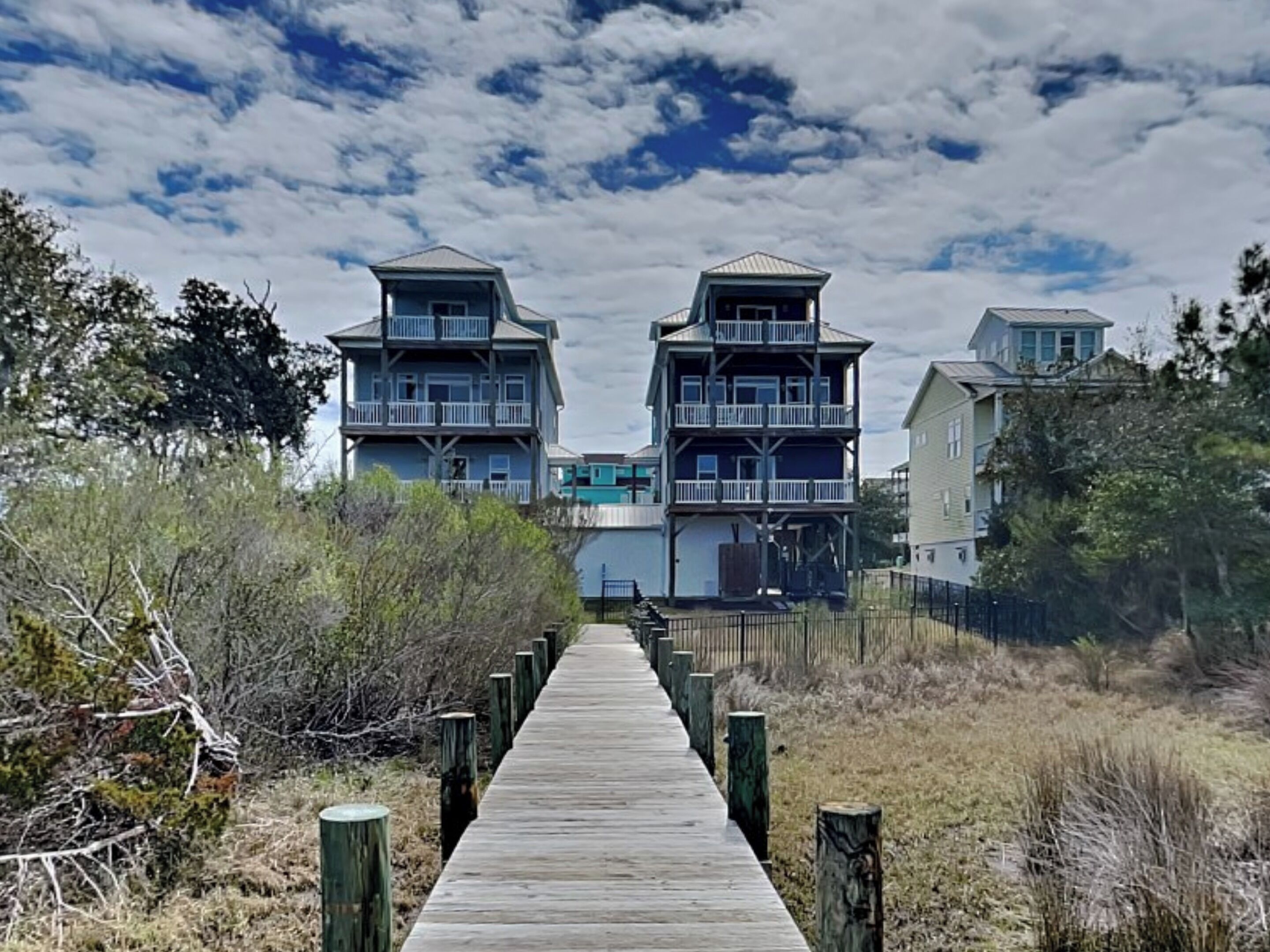 Une promenade en bois mène à une propriété située en bord de mer, avec des bâtiments à plusieurs étages, une zone clôturée et une végétation luxuriante tout autour.